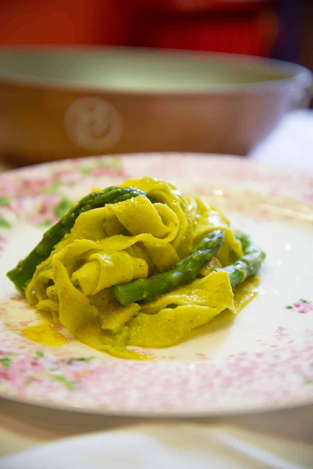 Piatto con decorazioni rosa. Tagliatelle con asparagi. Sullo sfondo una padella.