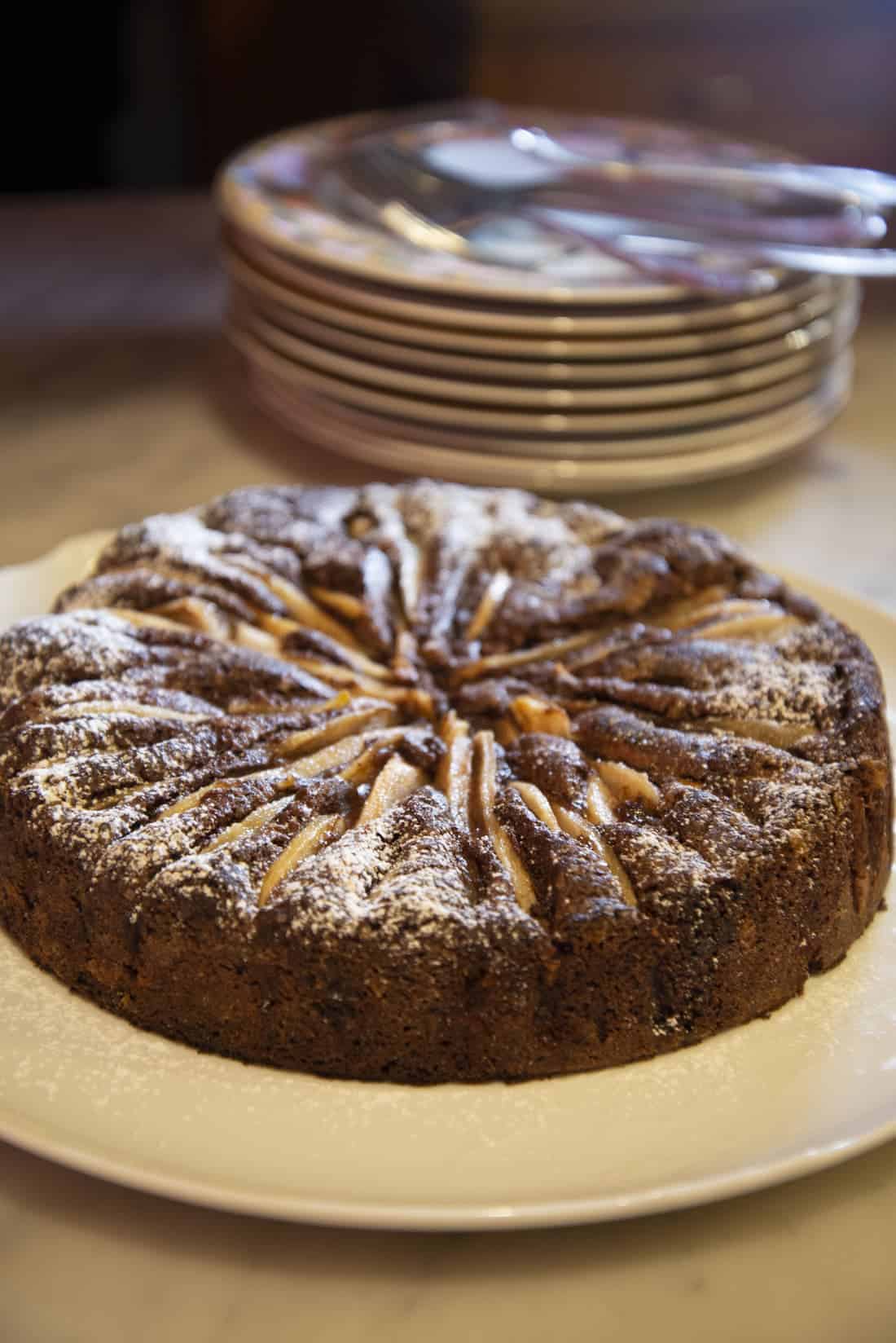 tavolo con sopra piatto bianco con torta al cioccolato, pere e zucchero. Piatti impilati sullo sfondo
